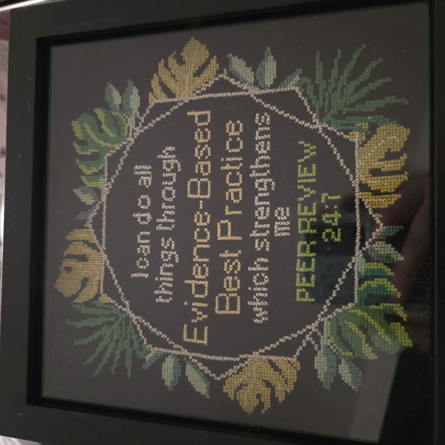 cross stitch of a wreath of leaves in different shades of green around a quote that reads "I can do all things through evidence based best practice which strengthens me. peer review 24:7"